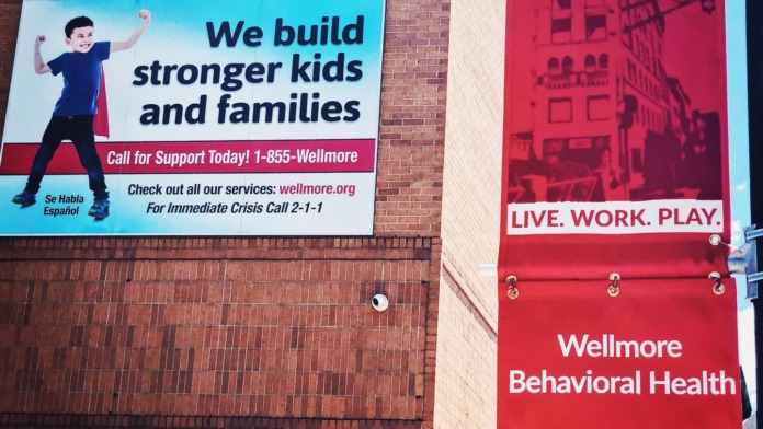 Exterior brick building wall displaying Wellmore Behavioral Health signage and marketing banner with facility contact information