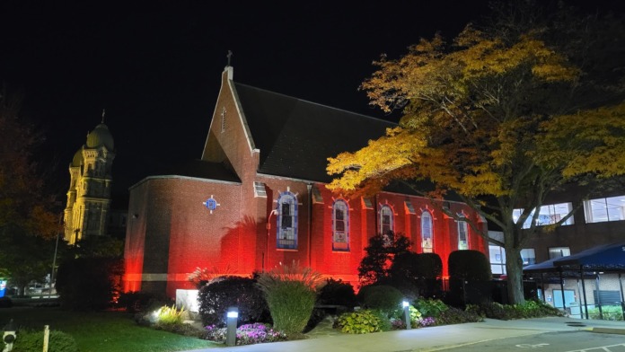 Red brick chapel building with red uplighting at night, landscaping and large tree in foreground