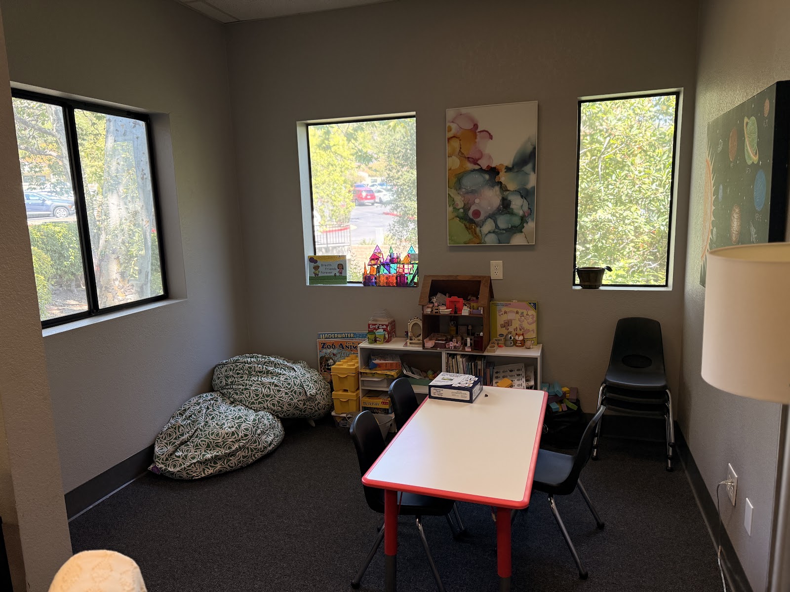 Interior children's activity room with colorful table, floor cushions, shelving with toys and books, and windows overlooking outdoor area