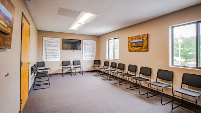 Interior meeting room with multiple black chairs in circle formation, cream colored walls, informational posters, television, large window with natural light, and wooden door