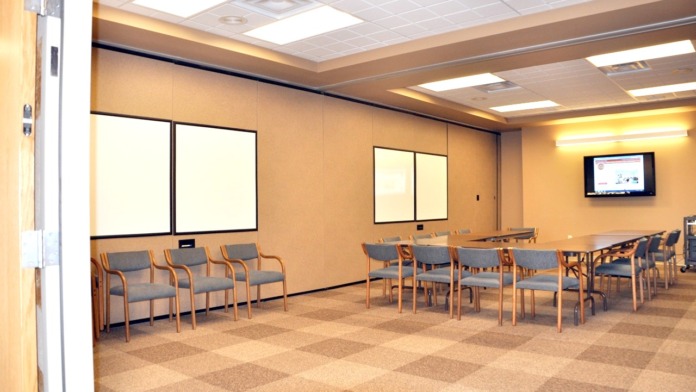 Interior meeting room with tan walls, classroom-style seating arrangement, whiteboard, and mounted television