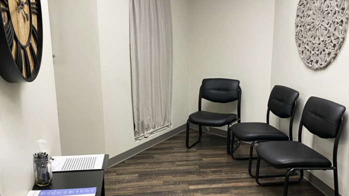 Interior waiting room with black client chairs, decorative wall art, clock, reception desk area with documents, and window with gray curtains.