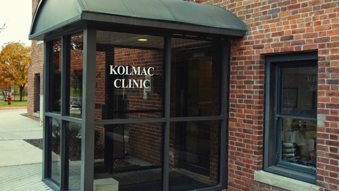 Exterior brick building entrance with dark green metal and glass canopy displaying KOLMAC CLINIC signage