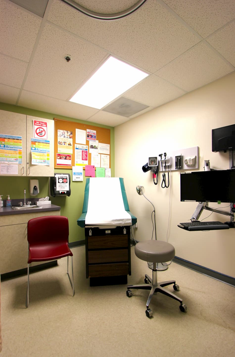 Interior clinical exam room with medical examination table, stool, medical equipment, and informational posters on walls