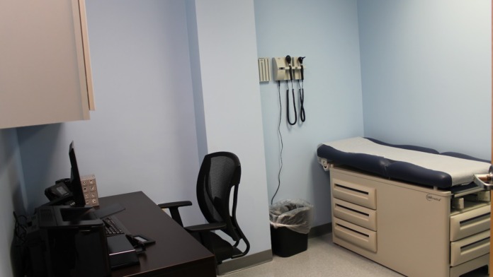 Interior clinical space with examination table, desk with computer workstation, and wall-mounted medical equipment.