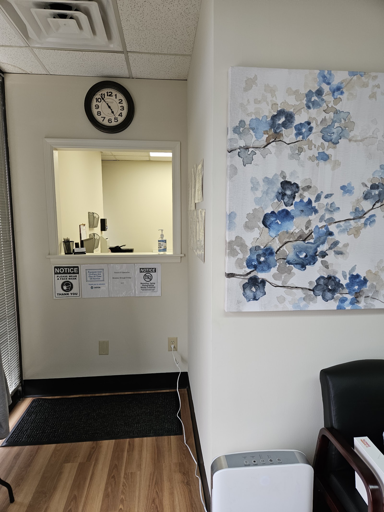 Interior office with reception window, wall clock, floral artwork, wooden flooring, and professional furnishings.