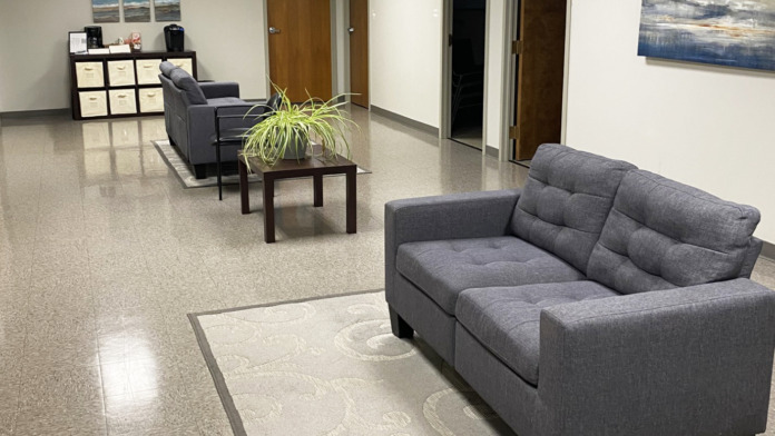 Interior common area with gray upholstered seating, dark flooring, interior doors, storage shelving, landscape artwork, and potted plant.