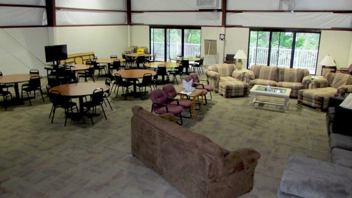 Interior common room with multiple round tables, upholstered seating, and windows overlooking exterior grounds.