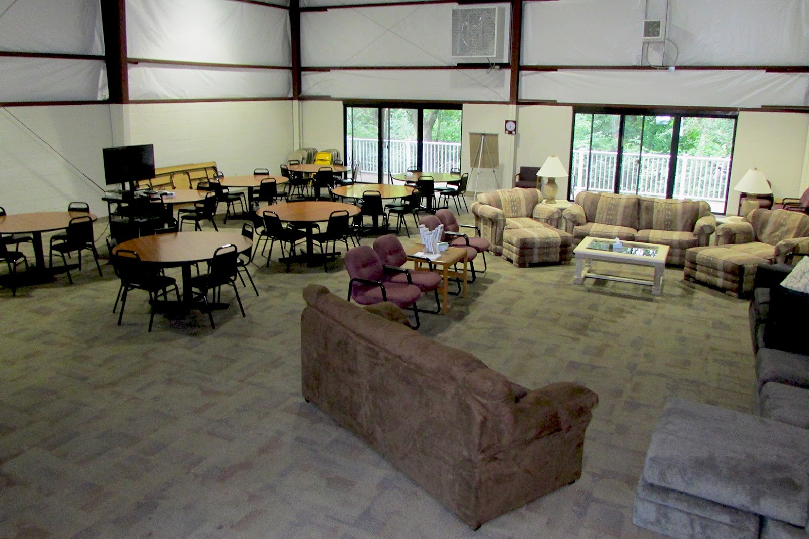 Interior common room with multiple round tables, upholstered seating, and windows overlooking exterior grounds.