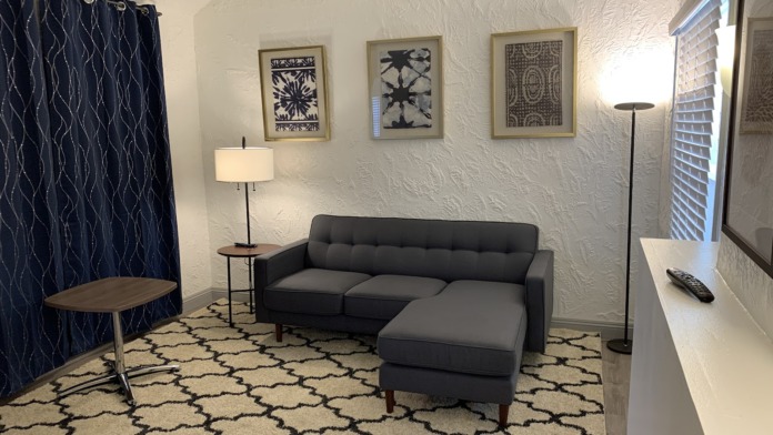 Interior lounge space with gray sectional sofa, patterned rug, wall art, table lamps, and window blinds