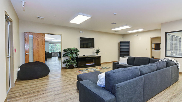 Interior common area with large gray sectional sofa, black leather chairs, fireplace, television, plant, and wood flooring