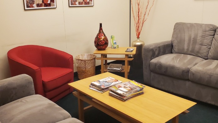 Interior common area with beige and gray upholstered chairs, yellow wooden side table with decorative vase, framed wall art, and magazines on tables