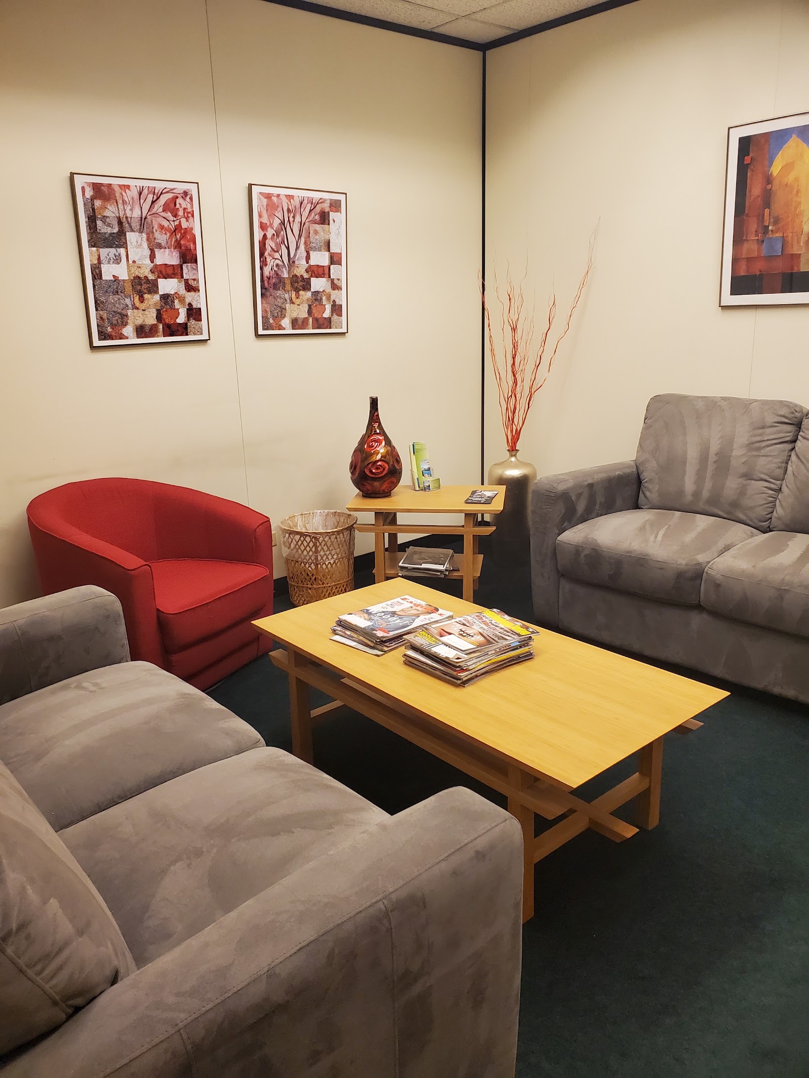 Interior common area with beige and gray upholstered chairs, yellow wooden side table with decorative vase, framed wall art, and magazines on tables