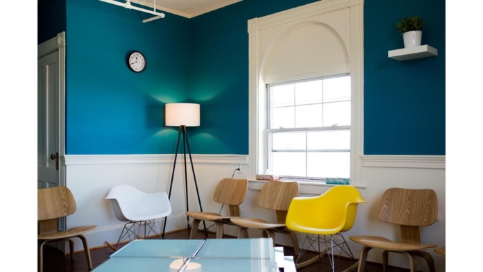 Interior common room with teal walls, white wainscoting, modern wood and colored chairs, floor lamp, wall clock, window with shade