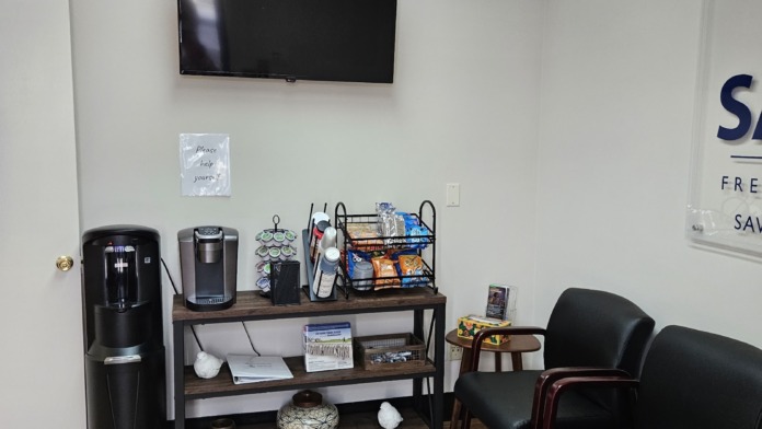 Interior break room with wall-mounted television, water cooler, black seating, refreshment station, and wooden flooring.