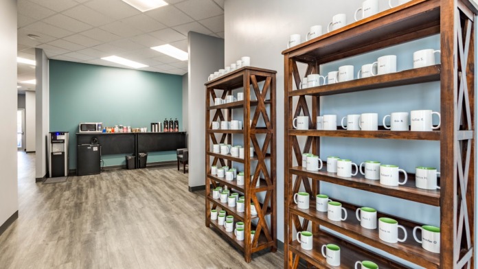 Interior break room with wooden shelving displaying personalized mugs, coffee equipment, teal accent wall, and beverage service area