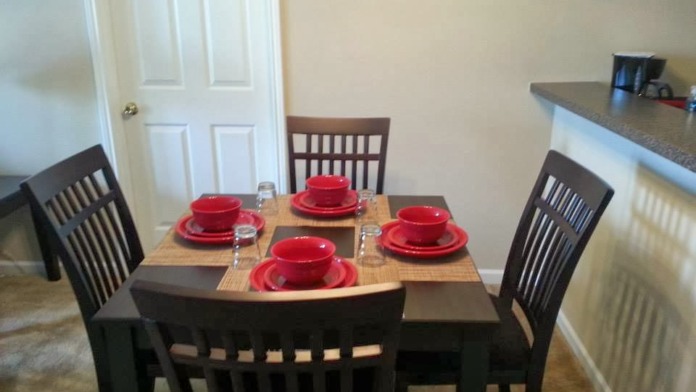 Interior dining area with round table, black chairs, red place settings, and kitchen counter visible in background