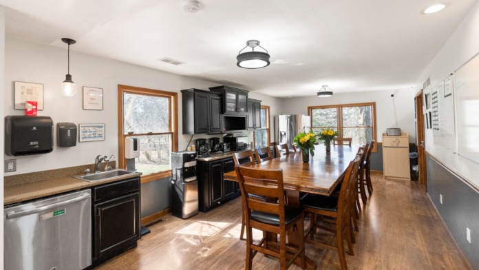 Interior dining room with large wooden table, seating for multiple people, kitchen with black cabinets, and natural light from windows