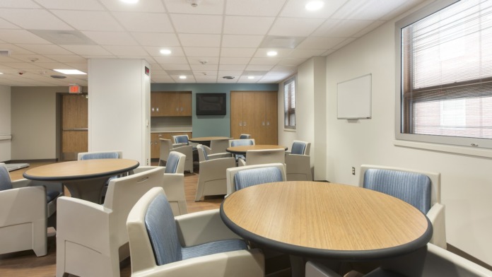 Interior common area with round tables, upholstered chairs, TV monitor, kitchenette, and natural lighting