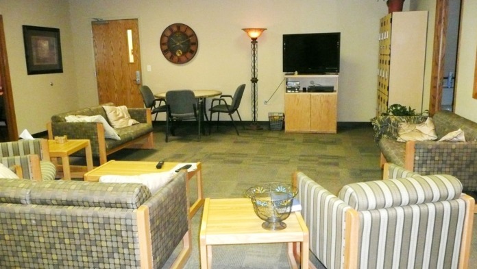 Interior residential common area with multiple seating arrangements, television, wall clock, potted plants, and warm lighting