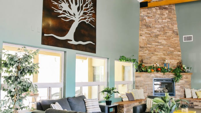 Interior living room with turquoise walls, gray sofa, fireplace with brick surround, large windows, tree artwork, and decorative plants