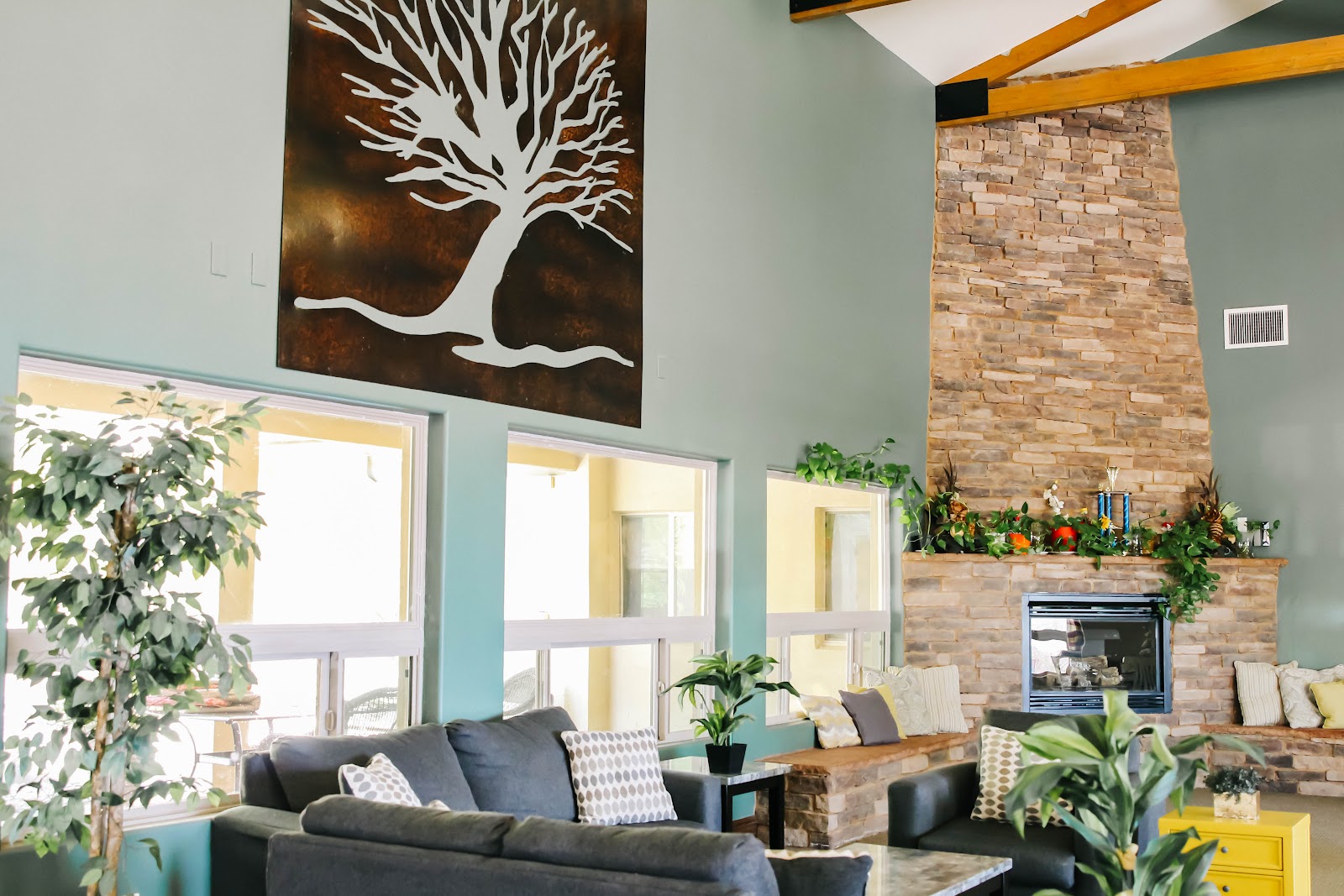 Interior living room with turquoise walls, gray sofa, fireplace with brick surround, large windows, tree artwork, and decorative plants