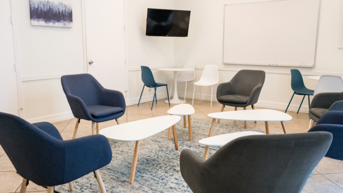 Interior community lounge with multiple comfortable chairs, tables, television, and natural lighting from large windows