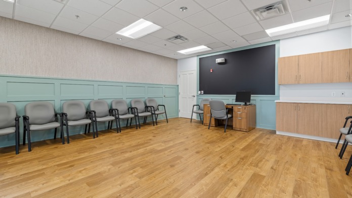 Interior community space with multiple gray chairs, green and white paneling, large display screen, wooden flooring, and storage cabinetry