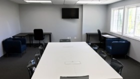 Interior conference room with long white table, black office chairs, wall-mounted television, and bright natural lighting from windows