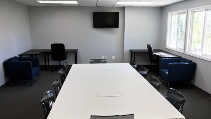 Interior conference room with long white table, black office chairs, wall-mounted television, and bright natural lighting from windows