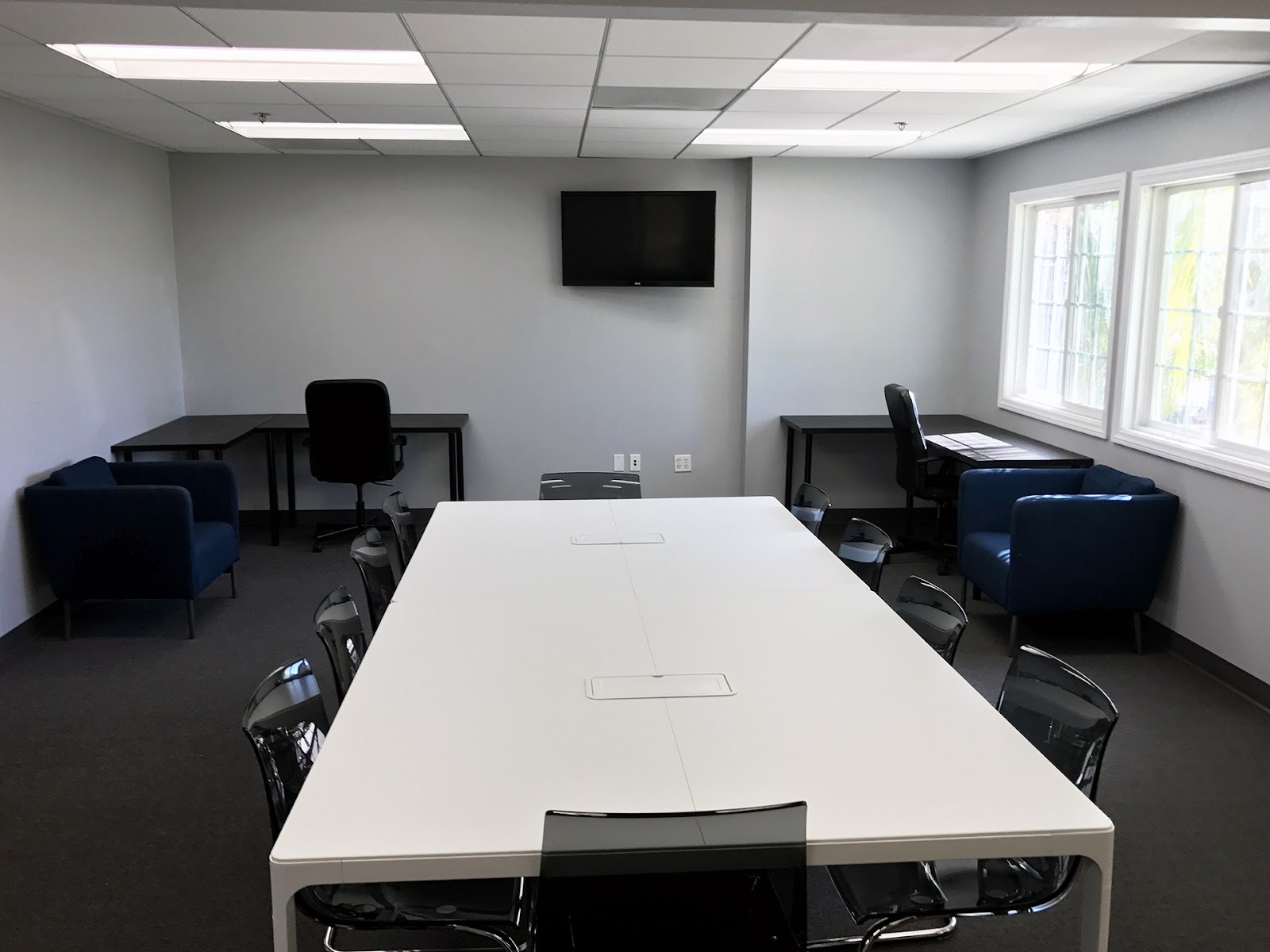 Interior conference room with long white table, black office chairs, wall-mounted television, and bright natural lighting from windows