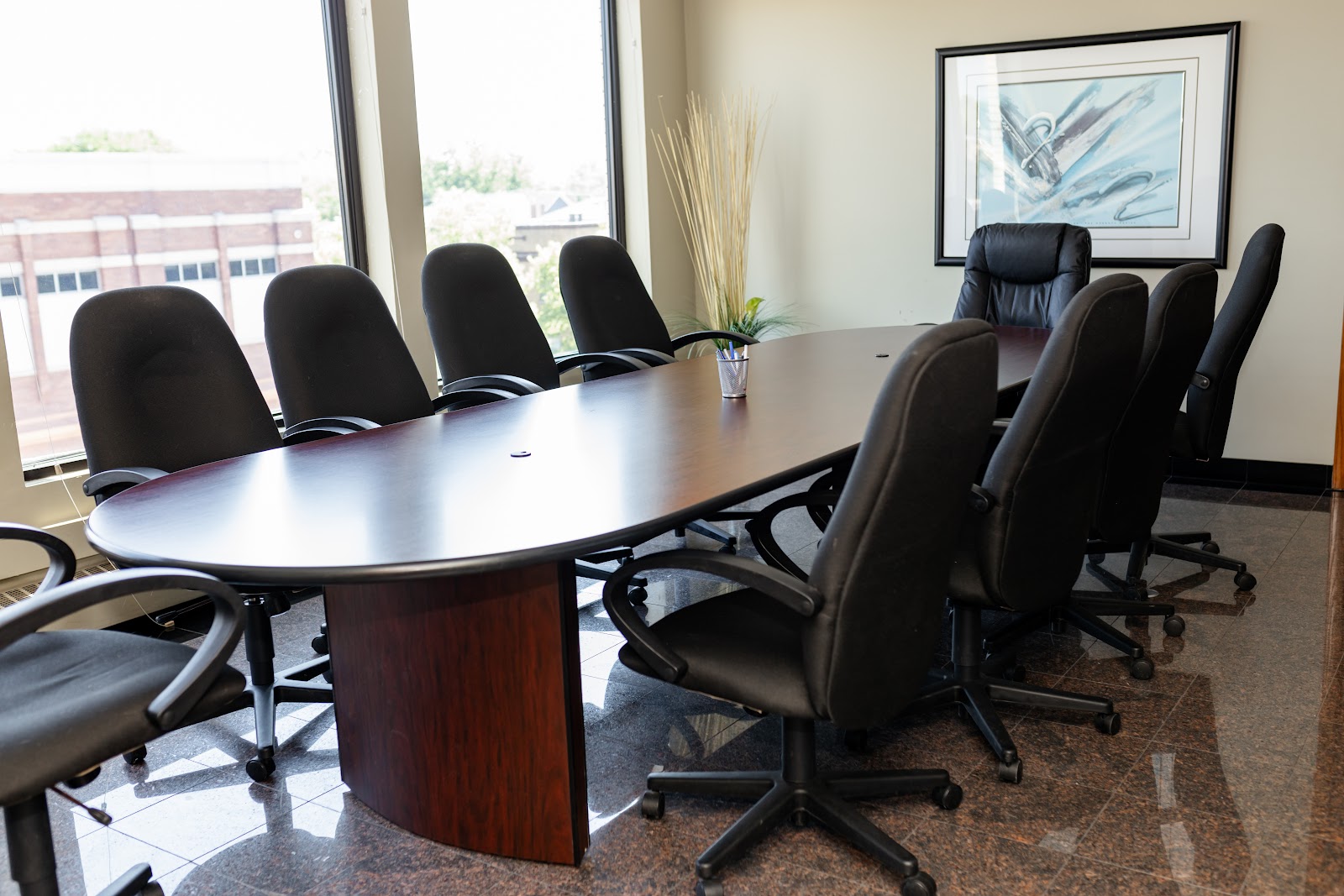 Professional conference room featuring oval wood table, executive black chairs, city views, and framed artwork