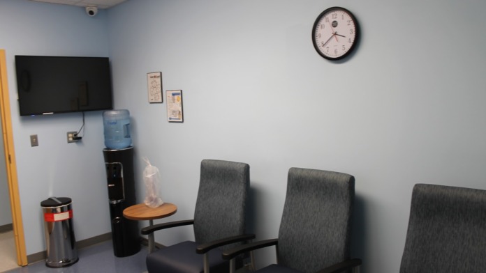 Interior waiting room with television display, water cooler, wall clock, and multiple upholstered chairs.