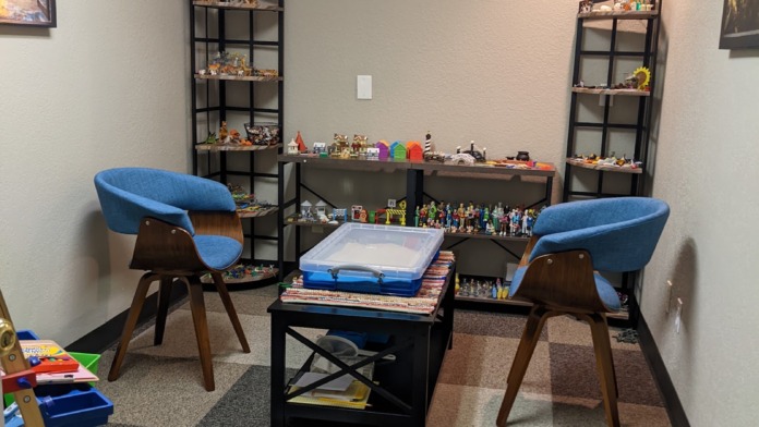 Interior therapy room with two modern blue chairs, black tables, shelving with decorative items, and framed wall artwork.