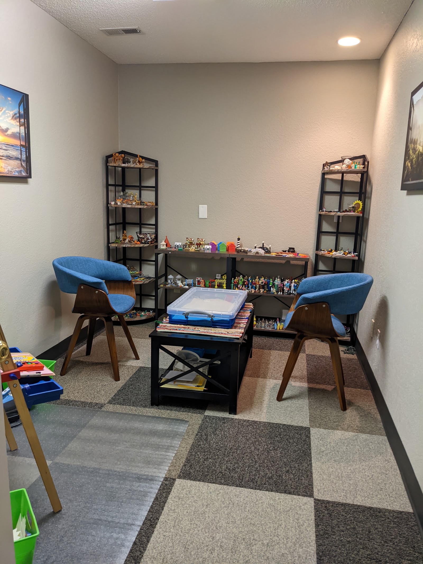 Interior therapy room with two modern blue chairs, black tables, shelving with decorative items, and framed wall artwork.