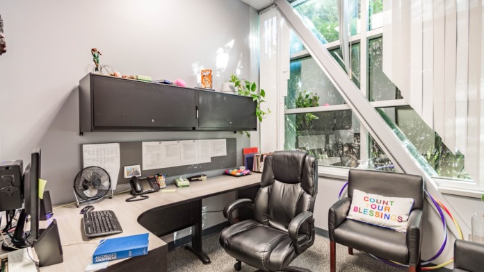 Interior office space with desk, black leather office chair, inspirational pillow, framed artwork, and large windows with tree views