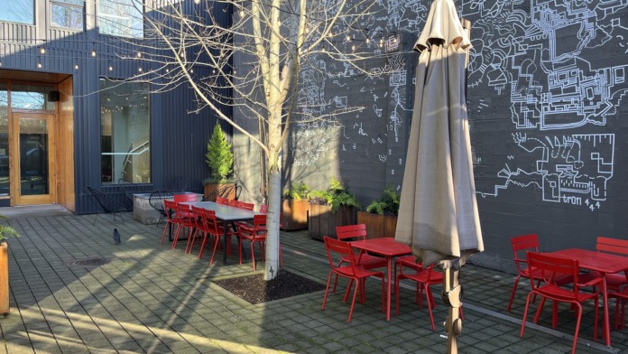 Outdoor patio featuring red metal chairs, white tables, bare tree, beige umbrella, and dark building wall with architectural drawings.