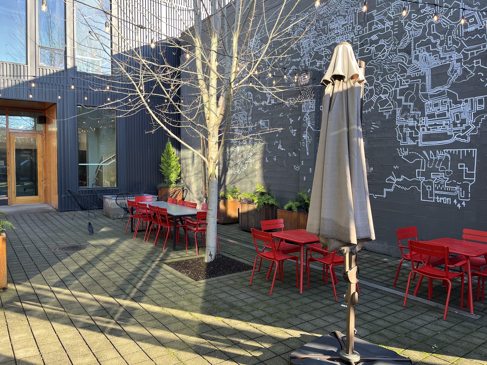 Outdoor patio featuring red metal chairs, white tables, bare tree, beige umbrella, and dark building wall with architectural drawings.