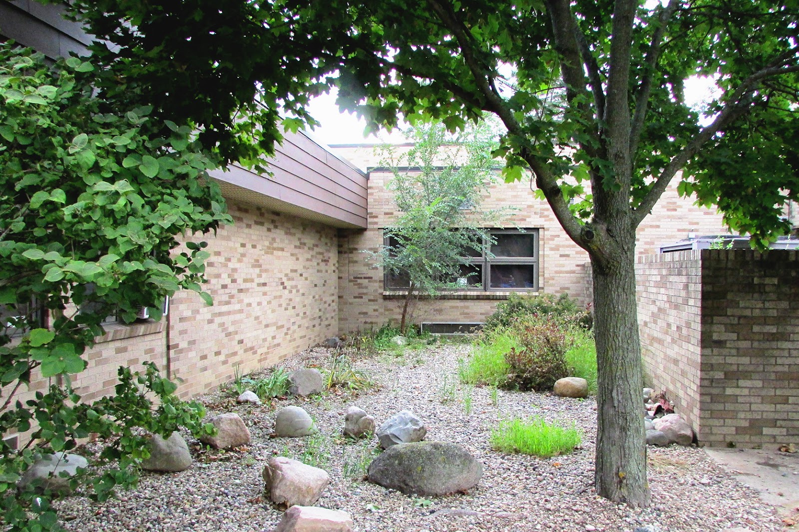 Exterior courtyard area with brick building, mature trees, gravel landscaping, and building windows.