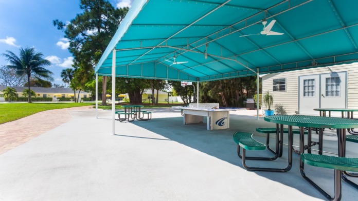 Exterior shaded pavilion with turquoise canopy, picnic tables, ping-pong table, yellow facility building visible in background, mature trees