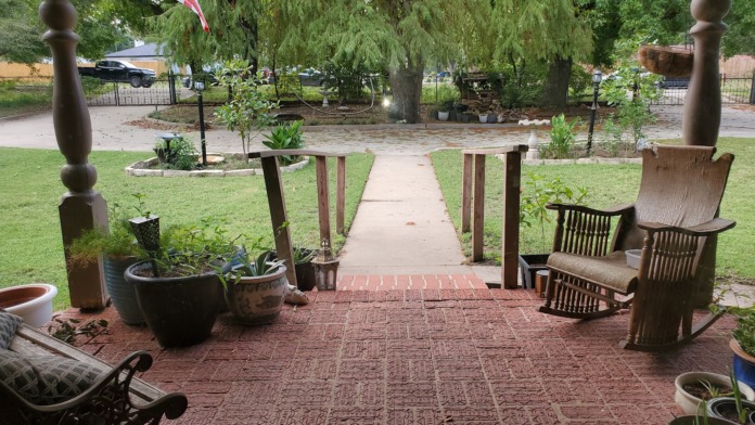 Covered patio with wooden posts, brick flooring, potted plants, Adirondack chairs, and flag