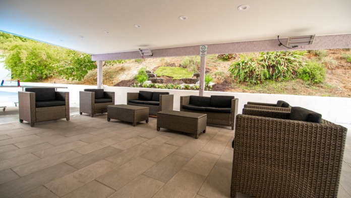 Outdoor covered patio with wicker furniture, black cushioned seating, potted plants, and hillside backdrop.