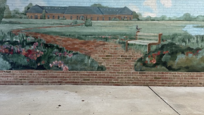 Interior wall mural depicting rural landscape with building, water, and flowering plants in watercolor style