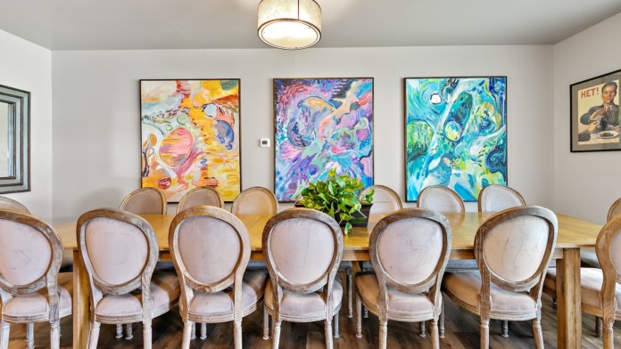 Interior dining space with long wooden table, upholstered chairs, vibrant abstract paintings, and decorative lighting