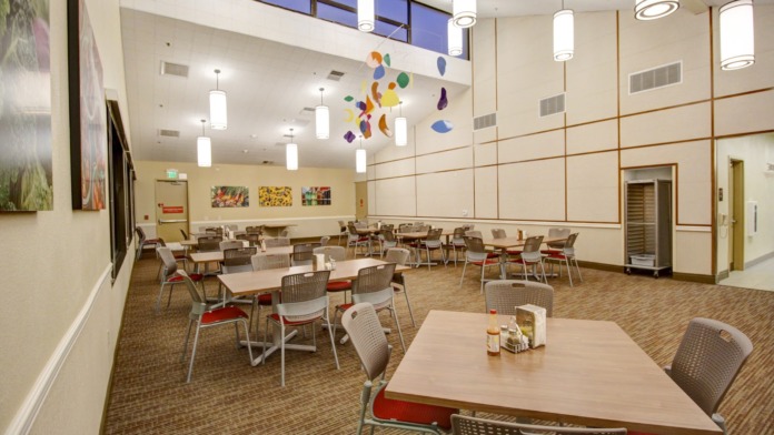 Interior multi-purpose room with round dining tables, chairs with red cushions, colorful ceiling mobile artwork, and modern pendant lighting