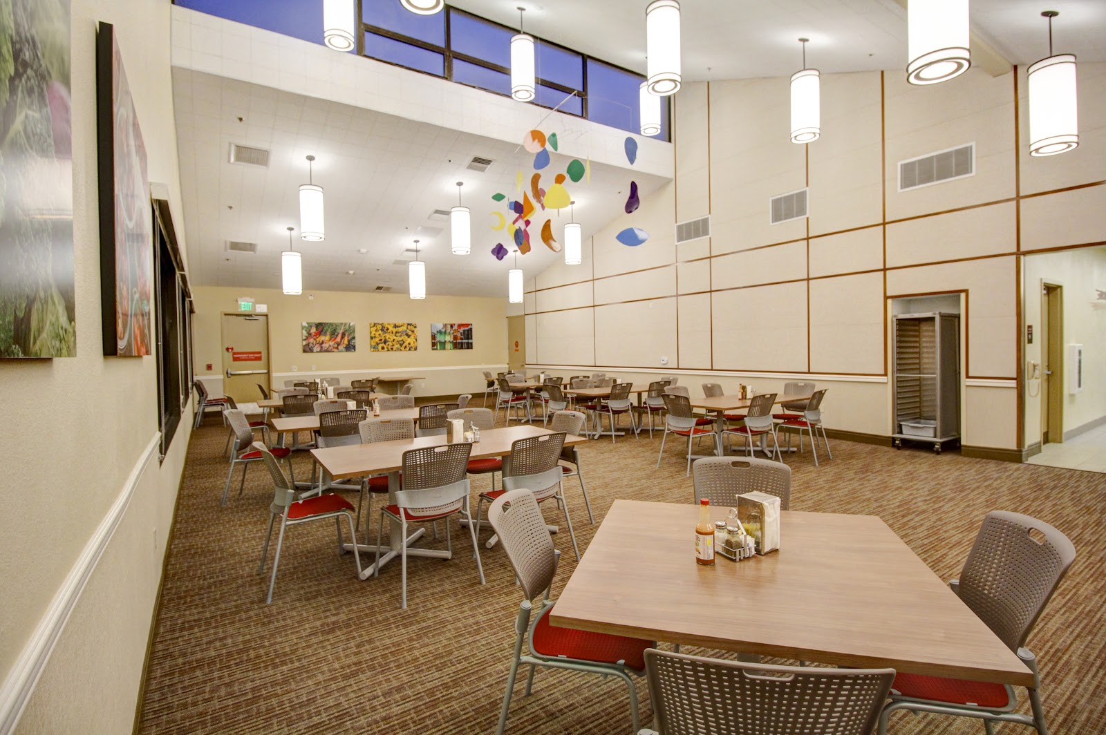 Interior multi-purpose room with round dining tables, chairs with red cushions, colorful ceiling mobile artwork, and modern pendant lighting