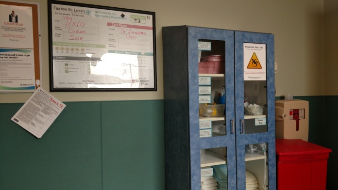 Interior healthcare facility room displaying Faxton St. Luke's signage, emergency department information board, storage cabinets, and safety signage