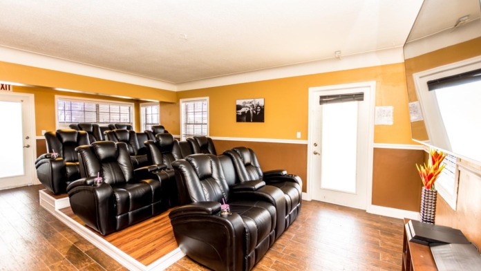Interior entertainment room with tiered black leather reclining chairs arranged in rows, orange and brown accent walls, and windows