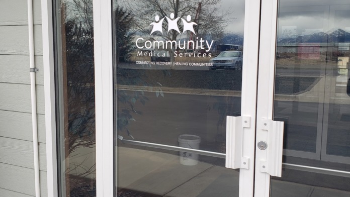 Exterior entrance doors with white frame and Community Medical Services logo visible, showing parking lot and landscape