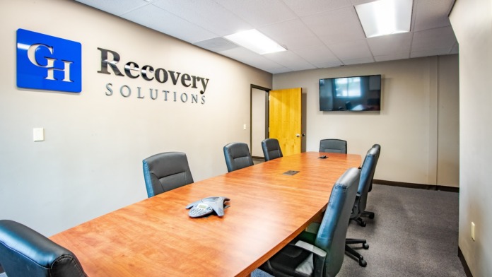 Interior conference room featuring large wooden table, blue and gray office chairs, facility logo on wall, television, and yellow door
